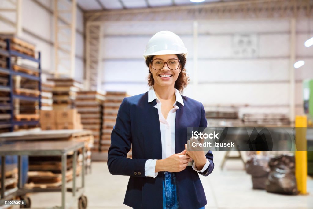 Portrait of smiling businesswoman wearing hardhat Portrait of smiling businesswoman wearing hardhat in industry. Confident inspector is on visit in warehouse. She is holding digital tablet. Business Stock Photo