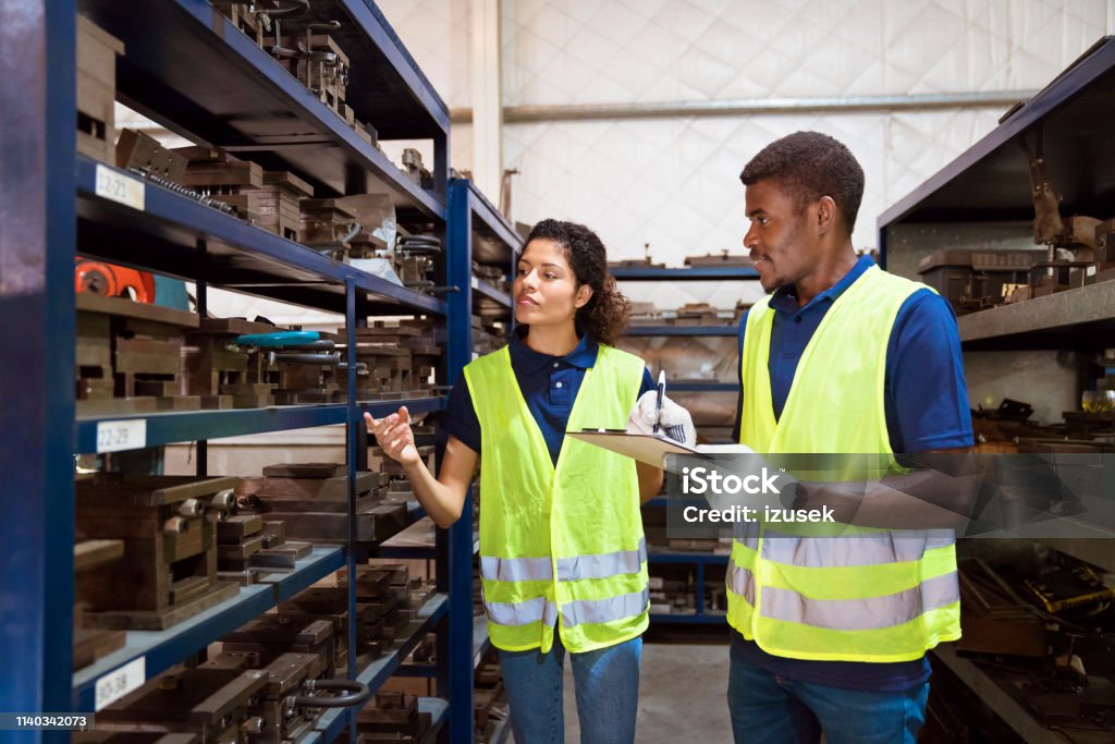 Production workers examining equipment on racks Production workers examining industrial equipment on racks. Expertise are wearing reflective clothing. Male engineer is holding clipboard by woman in factory. 25-29 Years Stock Photo