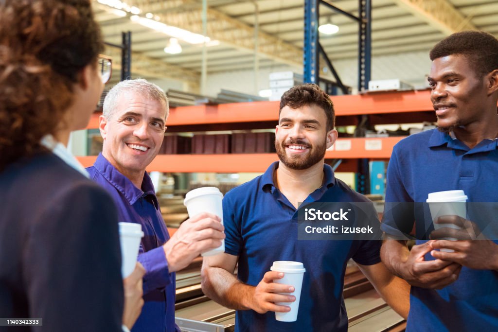 Engineers taking coffee break with female manager Engineers taking coffee break with manager in warehouse. Inspector is on visit in manufacturing industry. They are holding disposable cups. Coffee - Drink Stock Photo