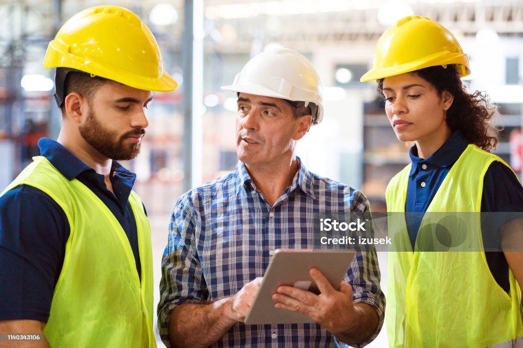 Man discussing over digital tablet with colleagues Foreman discussing over digital tablet with colleagues in factory. Engineers are working together in manufacturing company. They are wearing hardhats. Expertise Stock Photo