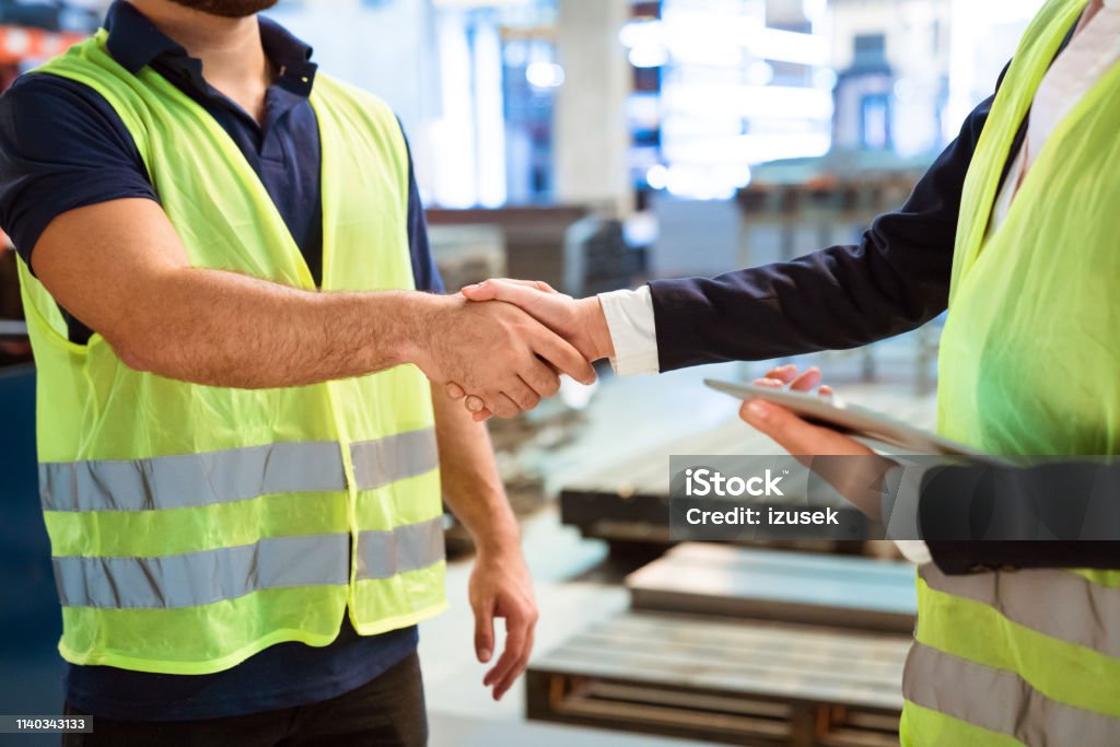 Manager and worker greeting each other in factory Midsection of manager shaking hand with production worker in factory. Engineers are wearing reflective clothing. They are greeting each other. Handshake Stock Photo