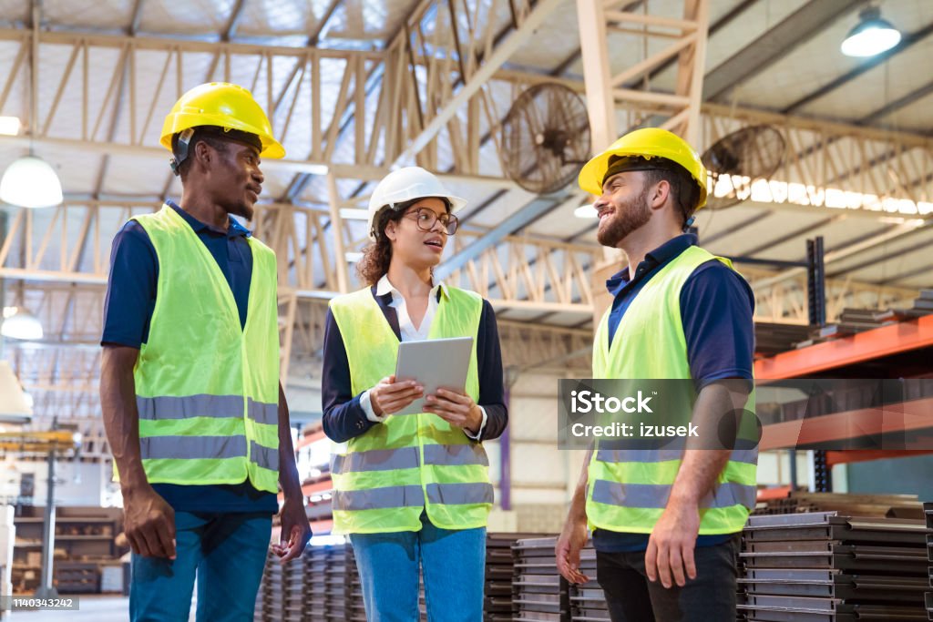 Manager with digital tablet talking to workers Female manager talking with production workers while holding digital tablet. Engineers are related to manufacturing occupation. They are wearing reflective clothing and hardhats in factory. 20-24 Years Stock Photo