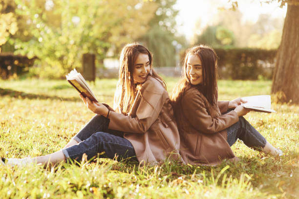 lächelnde brünette zwillingsmädchen, die sich hinten auf das gras setzen und sich gegenseitig betrachten, beine leicht in die knie gebogen, mit braunen büchern in den händen, das lässige fell im herbstpark auf blödem hintergrund - knees bent stock-fotos und bilder