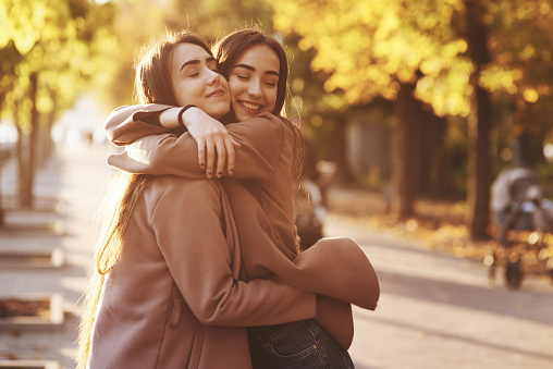 Perfil lateral de joven morena bonita sonrisa gemelas abrazándose y divirtiéndose en el abrigo informal de pie cerca el uno del otro en el soleado callejón del parque de otoño en el fondo borroso photo