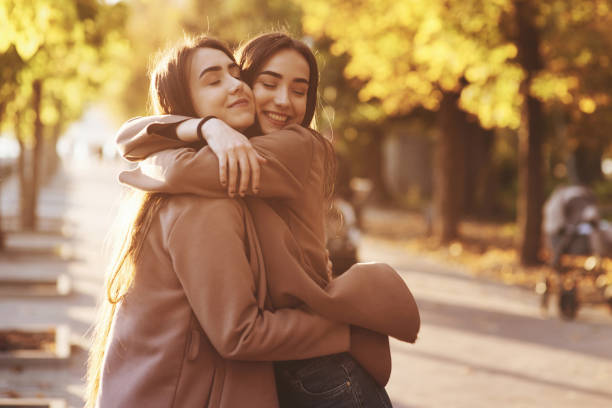 Side profile of young pretty smiling brunette twin girls hugging and having fun in casual coat standing close to each other at autumn sunny park alley on blurry background Side profile of young pretty smiling brunette twin girls hugging and having fun in casual coat standing close to each other at autumn sunny park alley on blurry background. sister stock pictures, royalty-free photos & images