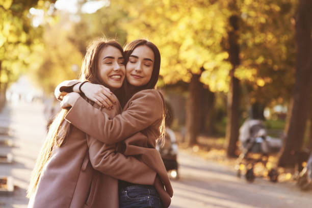 perfil lateral de meninas gêmeas triguenhas de sorriso novas que abraçam e que têm o divertimento no revestimento ocasional que está perto de se na aléia ensolarada do parque do outono no fundo obscuro - chearful - fotografias e filmes do acervo