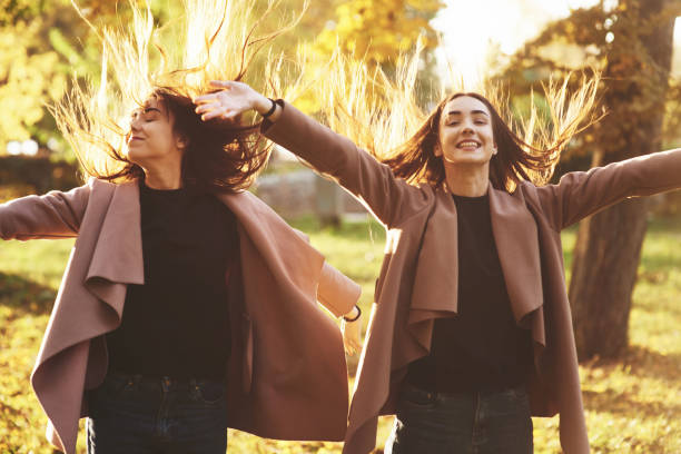 Young happy, smiling brunette twin girls standing and spreading hands wide open, their hair is dispelled up, wearing casual coat at autumn sunny park on the light blurry background. Freedom concept Young happy, smiling brunette twin girls standing and spreading hands wide open, their hair is dispelled up, wearing casual coat at autumn sunny park on the light blurry background. Freedom concept. dispelled stock pictures, royalty-free photos & images