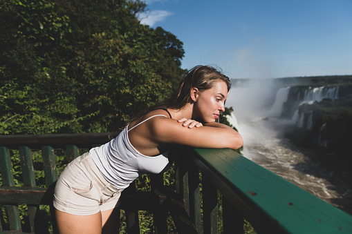 A young woman stands on a viewing platform on the Brazilian side of Iguazu Falls, looking contemplatively at the numerous waterfalls and lush landscape.