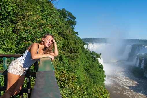 A young woman stands on a viewing platform on the Brazilian side of Iguazu Falls, smiling happily at the numerous waterfalls and lush landscape on the Argentinian side.