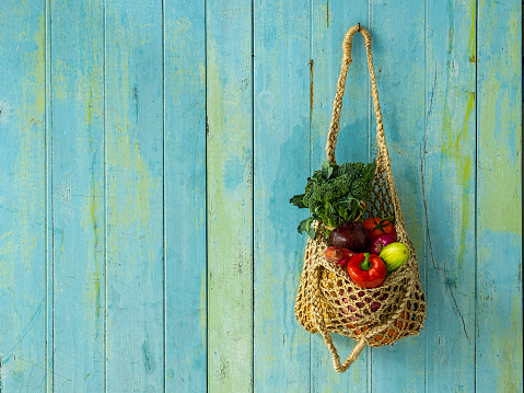 Many colorful contrast color salad vegetables in a reusable string jute bag hanging from an old blue and green abstract weathered wood panel background wall, with good copy space to the left of the image.