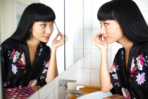 Chinese woman putting make up on, doing her eyebrows. Looking at mirror, side view. Close up
