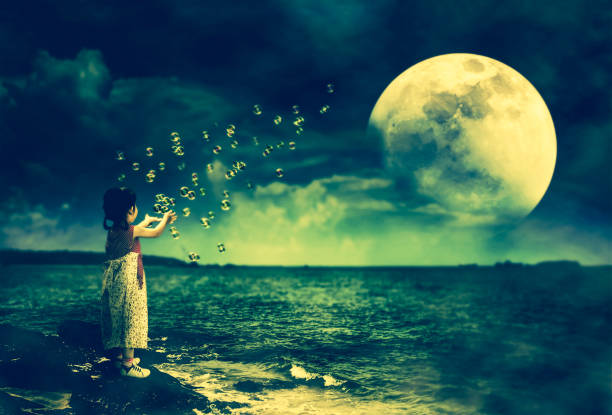 Back view of asian girl playing with soap bubbles on stone at seaside. Landscape of night sky with super moon behind partial cloud. Beautiful serenity background. The moon taken with my own camera.