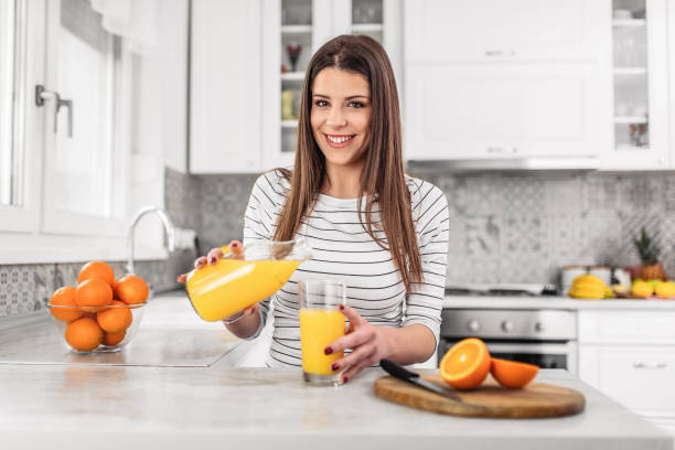 Close up portrait of a woman pouring orange juice in the glass while sitting at the kitchen stock photo