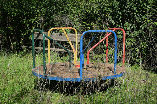 Old rusty playground merry-go-round and overgrown withered plants in abandoned park.