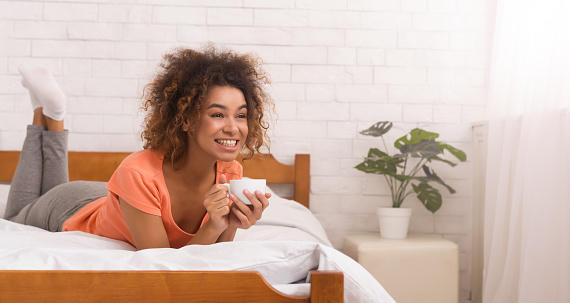 African-american girl relaxing in bed, enjoying and drinking coffee at morning, empty space