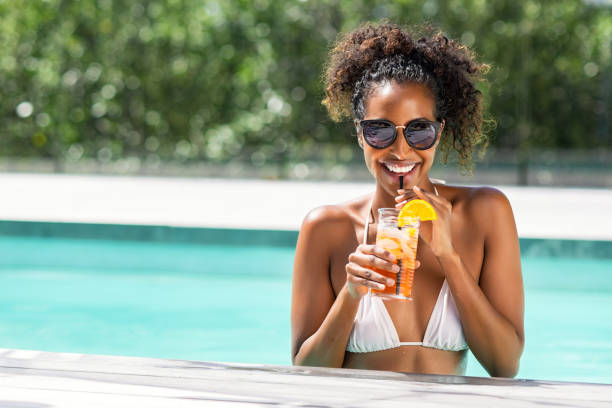 Fashion beauty woman in pool drinking cocktail Portrait of happy fashion woman with sunglasses standing in swimming pool drinking cocktail. Beautiful african glamour girl with fresh soft drink for appetizer in luxury pool looking at camera. Young woman with shades holding tumbler cocktail enjoying summer vacation. straw photos stock pictures, royalty-free photos & images