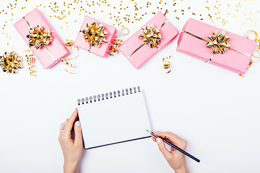 Female's hands writing in blank notepad near festive bright pink gift boxes with golden bows decorated with spangles and streamers on white background with copy space, view from above.