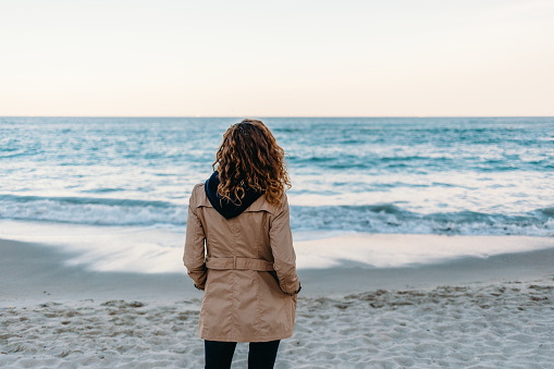 Rear view of young slender woman standing on beach looking at sea. Female with curly hair dressed in beige trench relaxing enjoying view alone on the sandy shore.