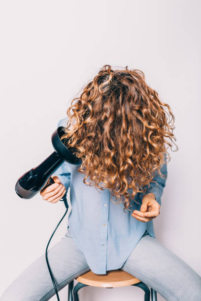 Young woman sitting on chair styling Young woman sitting on chair styling her curly hair with hairdryer with special diffuser nozzle. blow drying stock pictures, royalty-free photos & images