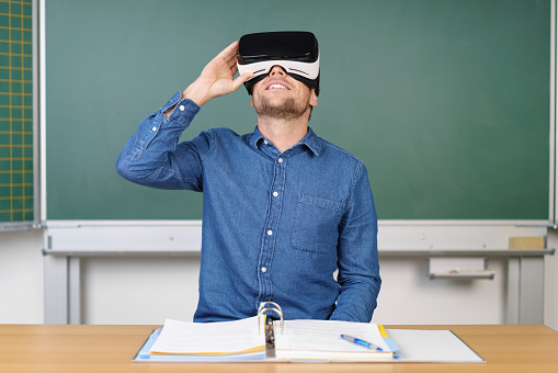 Young man looking up through 3D glasses VR headset sitting at teachers table against green chalkboard