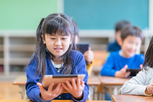 Elementary school children are using digital tablets and studying in a classroom.