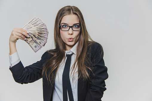 Satisfied, excited young business woman is showing a pile of money, isolated on white background. Girl has a lot of money. Cheerful girl is rounding her lips, because of astonishment of having money.