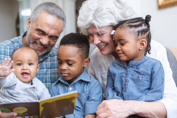 Story time with Grandma and Grandpa A retired senior couple support family by babysitting. Three young children sit on their grandparents' laps. The group is reading a book together. young at heart stock pictures, royalty-free photos & images
