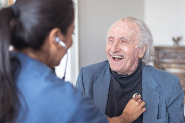 Happy and heathy! An elderly man smiles while meeting with his doctor. The female doctor is using a stethoscope to listen to the man's heart. The doctor has come to visit the man in his home. heathy stock pictures, royalty-free photos & images