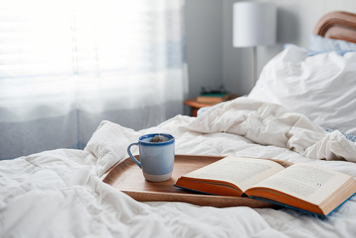 A steaming cup of hot tea and a book on wooden tray on unmade bed