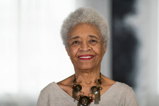 A senior woman of African descent is laughing at the camera while posing in front of a backdrop. She is elegant and poised.