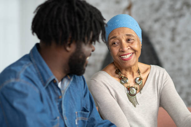 Senior Mother With Cancer Spending Time With Her Adult Son An African woman with cancer is happily spending time with her adult son in a living room. They are laughing and being affectionate. survival stock pictures, royalty-free photos & images