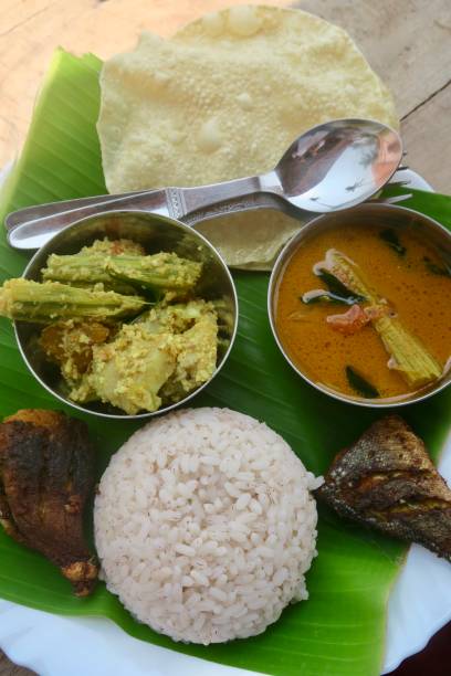 Photo showing traditional kerela, South Indian local food served on a green banana leaf as lunch or dinner. The food consists of papad / papadum / poppadom, fried king fish and spiced barbecued tuna steak, aviyal, alleppey coconut mango fish curry, matta / rosematta / Palakkadan with wild Keralan red rice mounded using an upside down bowl as a mould. imagen de kerela casera tradicional, comida local del sur de la india servida en una hoja de plátano. la comida consiste en papad/papadum/poppadom, pescado rey frito, aviyal, curry de pescado de coco con arroz rojo keralan silvestre montado usando un taz�