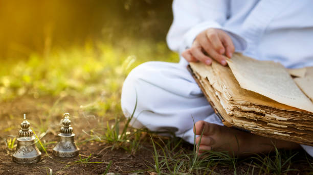 moine bouddhiste se trouve dans la position de lotus yoga méditant, lisant vieux livre ouvert de la sagesse, et il ya un nombre de cloches tibétaines et brûler l’encens au pré. beau fond de coucher du soleil - concentration contemplation clothing viewpoint photos et images de collection