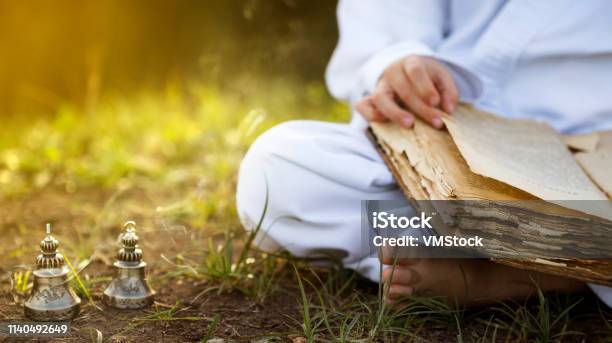 Buddhist Monk Sits In Yoga Lotus Position Meditating Reading Old Open Book Of Wisdom And There Are A Number Tibetan Bells And Burn Incense At Meadow Beautiful Sunset Background Stock Photo - Download Image Now