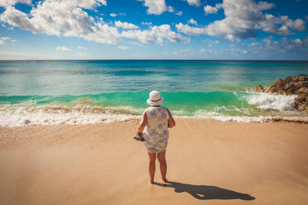 A middle aged woman in a dress and hat stands on a topical beach looking out to sea
