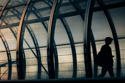 Color image depicting a back lit silhouette man walking by himself through an elevated glass walkway/tunnel in the city of London, UK. The architecture is futuristic and the glass is curved. The man appears to be a businessman or commuter. It is sunset with warm golden late afternoon light. Room for copy space.