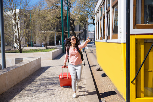 Young woman running after trolleybus and pulling trolley case. Pretty lady with spring city view in background. Tourism and transportation concept. Front view.