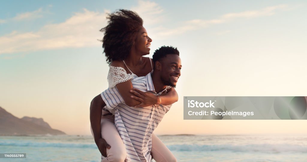 Journeying beyond the norm with my love Shot of a cheerful young woman being carried piggyback by her boyfriend at the beach Couple - Relationship Stock Photo