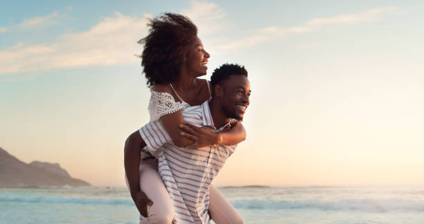 Journeying beyond the norm with my love Shot of a cheerful young woman being carried piggyback by her boyfriend at the beach black couple sunset stock pictures, royalty-free photos & images