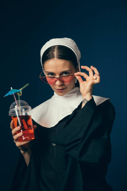 Medieval young woman as a nun Medieval bar and summertime. Young woman as a nun in vintage clothing and white mutch sitting on the chair on dark blue background. Drinking lemonad or coctail. Concept of comparison of eras. young nun stock pictures, royalty-free photos & images