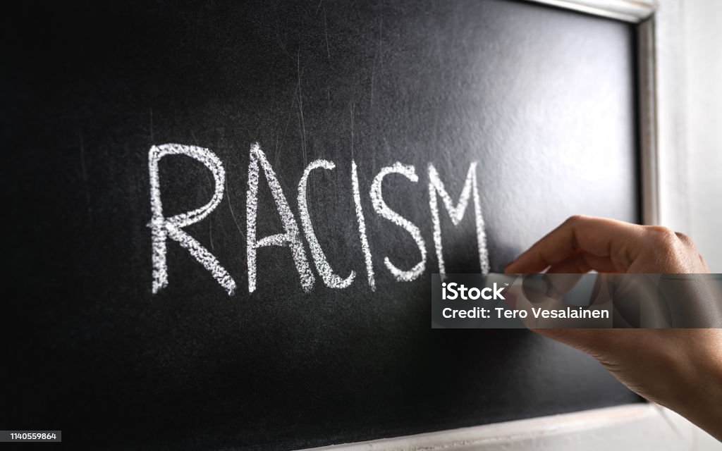 Hand writing the word racism on blackboard. Stop hate. Against prejudice and violence. Lecture about discrimination in school. Hand writing the word racism on blackboard. Stop hate. Against prejudice and violence. Lecture about discrimination or stereotypes in school. Racism Stock Photo