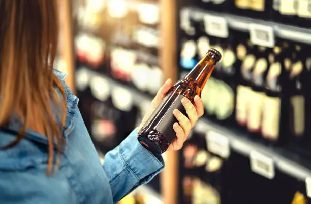 Photo of Customer buying beer in liquor store. Lager, craft or wheat beer. IPA or pale ale. Woman at alcohol shelf. Drink section and aisle in supermarket. Lady holding bottle in hand. Drink business.