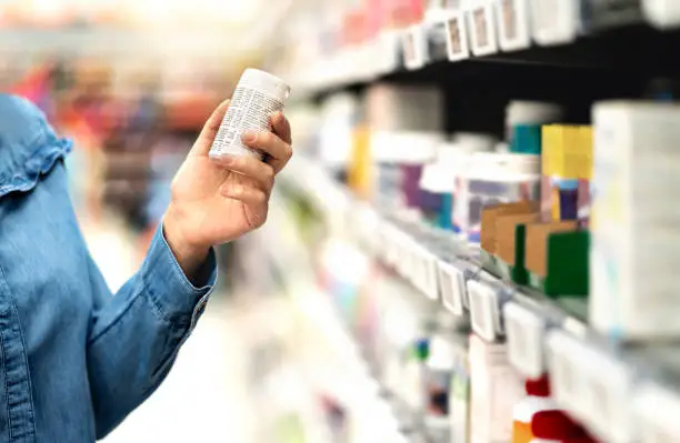 Photo of Customer in pharmacy holding medicine bottle. Woman reading the label text about medical information or side effects in drug store. Patient shopping pills for migraine or flu.