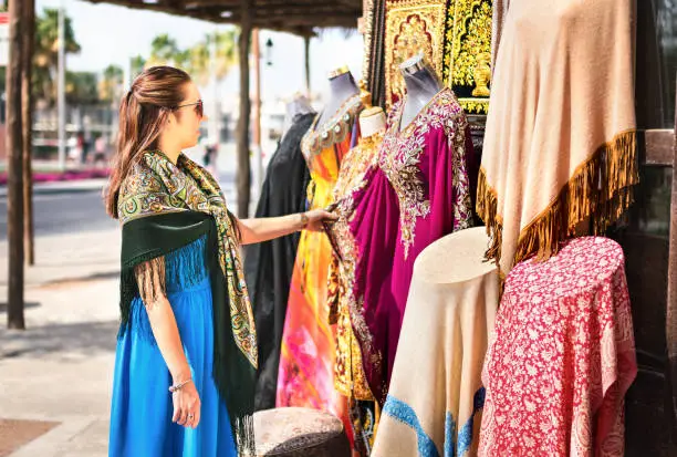 Woman in souk. Tourist looking at traditional Arabian dresses and clothes in store or outdoor market. Lady customer shopping souvenir in old Dubai. Tourism and vacation in the United Arab Emirates.