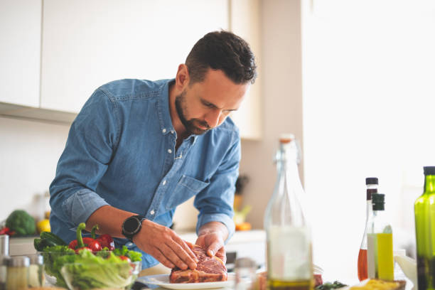 Bearded young man cooking meat while standing near kitchen table