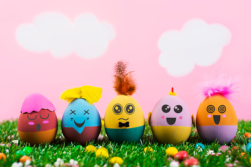 closeup of some handmade easter eggs, with cute and funny faces and hairstyles, on the grass with a pink sky with white clouds in the background