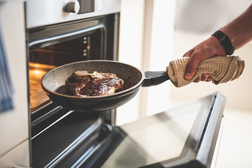 Close up of male hand holding frying pan with delicious beef steak near electric oven