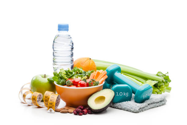 Healthy food and fitness concept: Front view of a bowl filled with fresh organic vegetables salad isolated on white background. Fruits and vegetables like green apple, orange, bell pepper, lettuce, celery, asparagus, avocado, cherry tomatoes and a water bottle are all around the salad bowl. A yellow and white tape measure and a pair of dumbbell weighs on a towel complete the composition. High key DSRL studio photo taken with Canon EOS 5D Mk II and Canon EF 100mm f/2.8L Macro IS USM.