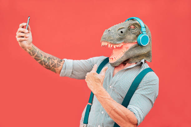 homem sênior louco que desgasta a máscara do t-rex e que toma o selfie com smartphone móvel-hipster macho mais idoso que tem o divertimento que escuta a música e a dança ao ar livre-conceitos absurdos, engraçados e surreal - body adornment - fotografias e filmes do acervo