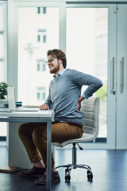 This is not good for my back Shot of a young businessman experiencing back pain while working at his desk in a modern office office back pain stock pictures, royalty-free photos & images